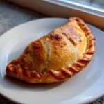 Nahaufnahme einer gebackenen Empanada mit perfekt gebr&auml;untem Teig auf einem wei&szlig;en Teller.