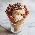 Nahaufnahme eines Eiswaffelh&ouml;rnchens mit zwei Kugeln Vanilleeis, betr&auml;ufelt mit Karamell- und Schokoladensauce und bestreut mit Schokost&uuml;ckchen.