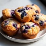Nahaufnahme von saftigen Mini Blaubeermuffins auf einem Teller, mit sichtbaren Blaubeeren und goldbrauner Kruste.