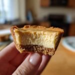 Eine Hand h&auml;lt einen k&ouml;stlichen Mini-K&auml;sekuchen mit einem Bissen, der den cremigen Belag und den knusprigen Boden zeigt.