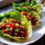 Nahaufnahme von drei Chicken Lettuce Wraps gef&uuml;llt mit gew&uuml;rztem Hackfleisch, roten Chilis und Fr&uuml;hlingszwiebeln auf einem wei&szlig;en Teller.