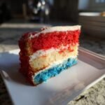 Ein St&uuml;ck Feuerwerk Kuchen mit roten, wei&szlig;en und blauen Schichten, garniert mit wei&szlig;er Glasur und roten Streuseln.