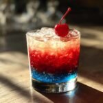 Ein erfrischender gefrorener Bomb Pop Cocktail mit roten und blauen Schichten, garniert mit einer Kirsche und viel Eis.