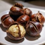 Nahaufnahme von mehreren perfekt ger&ouml;steten Kastanien auf einem wei&szlig;en Teller, eine davon ist bereits ge&ouml;ffnet und zeigt das weiche Innere.