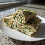 Vier angeschnittene Pinwheel Sandwiches mit Frischk&auml;se und Kr&auml;utern auf einem wei&szlig;en Teller.