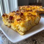 Ein St&uuml;ck Bacon And Eggs Breakfast Bake mit geschmolzenem K&auml;se und knusprigem Speck auf einem wei&szlig;en Teller.