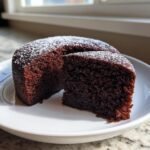 Ein St&uuml;ck saftiger Black Magic Chocolate Cake, best&auml;ubt mit Puderzucker, auf einem wei&szlig;en Teller.