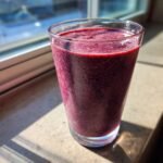 Ein hohes Glas mit einem tiefvioletten Blueberry Pomegranate Smoothie steht im Sonnenlicht auf einer Fensterbank.