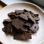 Nahaufnahme von zerbrochenen St&uuml;cken dunkler Chocolate Crackers, bestreut mit Kakaopulver, auf einem hellen Teller.