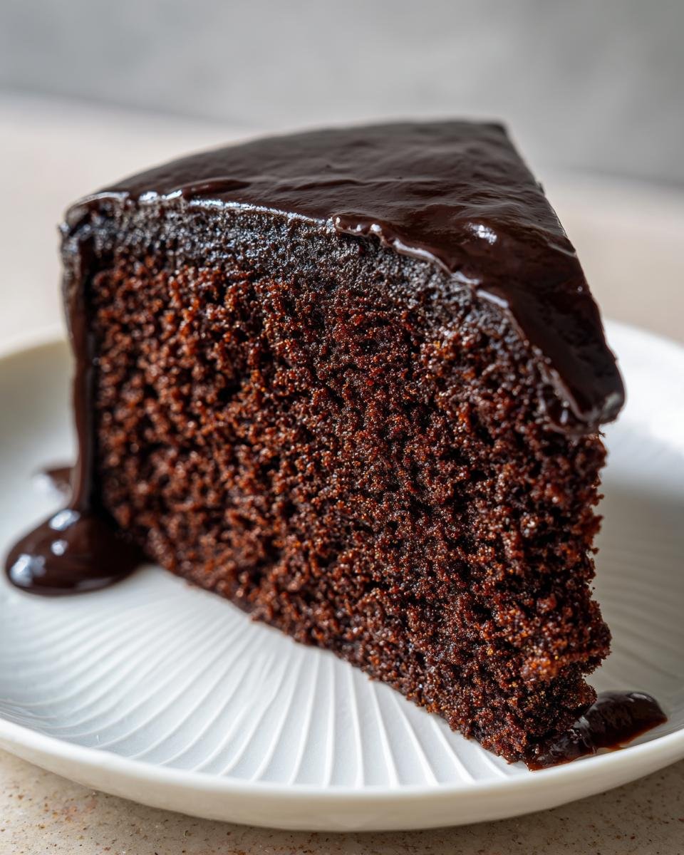 Nahaufnahme eines St&uuml;cks Chocolate Fudge Cake mit dunkler, gl&auml;nzender Schokoladenglasur auf einem wei&szlig;en Teller.