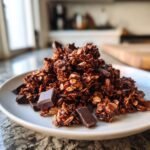 Ein Haufen knuspriges Chocolate Granola mit dunklen Schokoladenst&uuml;cken auf einem wei&szlig;en Teller.