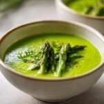 Nahaufnahme einer leuchtend gr&uuml;nen Asparagus Soup, garniert mit ganzen Spargelspitzen und Kr&auml;utern.