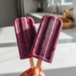 Nahaufnahme von zwei selbstgemachten Cremigen Blaubeer-Eis am Stiel, die in der Hand gehalten werden.