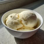 Zwei Kugeln cremiges, hellgelbes Homemade Ice Cream in einer wei&szlig;en Sch&uuml;ssel, beleuchtet von Sonnenlicht.