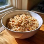 Eine wei&szlig;e Sch&uuml;ssel gef&uuml;llt mit leckerem Mandel-Naturreis-Pilaw, beleuchtet durch Sonnenlicht am Fenster.