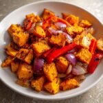 Nahaufnahme von gew&uuml;rfeltem, karamellisiertem Sweet Potato Hash mit roten Zwiebeln und Paprikastreifen in einer wei&szlig;en Sch&uuml;ssel.