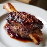 Nahaufnahme eines saftigen Wine Braised Lamb Shank mit dunkler, gl&auml;nzender So&szlig;e auf einem wei&szlig;en Teller.