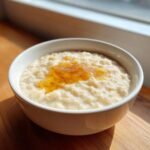 Eine wei&szlig;e Sch&uuml;ssel mit cremigem Porridge, garniert mit einem L&ouml;ffel goldgelbem Honig. Perfekt f&uuml;r Porridge Recipes.