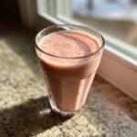 Ein Glas cremiger, rosafarbener Strawberry Cheesecake Protein Shake steht auf einer Granit-Arbeitsplatte.