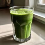 Ein Glas leuchtend gr&uuml;ner Green Kale Smoothie steht im Sonnenlicht auf einer hellen Arbeitsfl&auml;che.