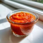 Nahaufnahme einer leuchtend orange-roten, dickfl&uuml;ssigen Hot Sauce in einem kleinen Glas.