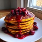 Ein hoher Stapel goldgelber K&uuml;rbispfannkuchen, &uuml;bergossen mit leuchtend rotem Preiselbeer-Ahornsirup und ganzen Cranberries.