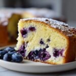 Nahaufnahme eines St&uuml;cks Lemon Blueberry Cream Cheese Pound Cake mit Blaubeeren und Puderzucker.