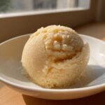 Eine Kugel cremiges, hellgelbes Marzipan-Eis in einer wei&szlig;en Schale, beleuchtet von Sonnenlicht.