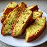 Nahaufnahme von goldbraun gebackenen Scheiben Pistazien Biscotti, reich an gr&uuml;nen Pistazienst&uuml;cken, auf einem wei&szlig;en Teller.