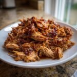 Nahaufnahme von zartem, gezupftem Schweinefleisch (Pulled Pork) mit dunkler Glasur, serviert auf einem wei&szlig;en Teller.