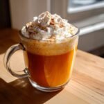 Nahaufnahme eines leuchtend orangefarbenen Pumpkin Spice Latte in einer Glastasse mit viel Schlagsahne und Zimt.
