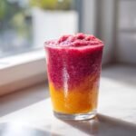 Ein Glas mit einem zweischichtigen, gefrorenen Raspberry Mango Smoothie, unten orange und oben lila-rot.