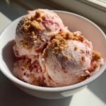 Nahaufnahme von drei Kugeln Strawberry Cheesecake Ice Cream mit Keksst&uuml;ckchen in einer wei&szlig;en Schale.