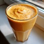 Ein cremiger, orangefarbener Sweet Potato Pie Smoothie mit Schaumkrone in einem Glas, das im Sonnenlicht steht.