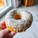 Eine Hand h&auml;lt einen gebackenen Vegan Lemon Poppyseed Donut mit dickem, wei&szlig;em Zitronenguss und Mohnsamen.