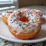 Nahaufnahme eines glasierten, veganen Vanille Donuts mit bunten Streuseln auf einem wei&szlig;en Teller.