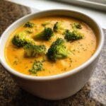 Nahaufnahme einer cremigen, orangefarbenen Broccoli Cheddar Suppe mit gro&szlig;en Brokkolir&ouml;schen in einer wei&szlig;en Sch&uuml;ssel.