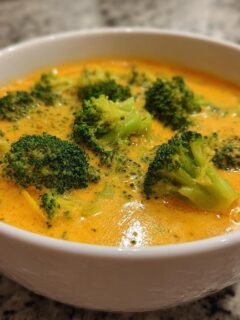 Nahaufnahme einer cremigen, gelb-orangenen Broccoli Cheddar Suppe mit großen Brokkoliröschen in einer weißen Schüssel.
