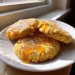 Drei Crumbl Cornbread Cookies auf einem wei&szlig;en Teller, betr&auml;ufelt mit gl&auml;nzendem Honig und Zucker.