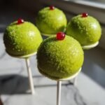Vier leuchtend gr&uuml;ne Grinch Cake Pops mit Zuckerstreuseln und einer roten Kirsche obenauf, die im Gegenlicht stehen.