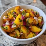 Nahaufnahme einer wei&szlig;en Schale gef&uuml;llt mit leuchtend gelber Peach Salsa, gew&uuml;rfeltem Pfirsich, roten Zwiebeln und Kr&auml;utern.