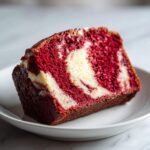 Nahaufnahme eines saftigen St&uuml;cks Red Velvet Banana Bread mit deutlicher roter und wei&szlig;er Marmorierung auf einem wei&szlig;en Teller.