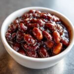 Nahaufnahme von gl&auml;nzenden, dunkelbraunen Root Beer Baked Beans in einer kleinen wei&szlig;en Sch&uuml;ssel.