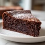 Nahaufnahme eines saftigen St&uuml;cks Flourless Chocolate Cake, best&auml;ubt mit Puderzucker auf einem wei&szlig;en Teller.