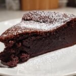 Ein saftiges St&uuml;ck Flourless Chocolate Cake, best&auml;ubt mit Puderzucker, auf einem wei&szlig;en Teller.