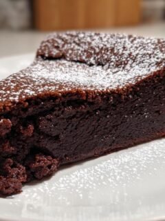 Ein saftiges Stück Flourless Chocolate Cake, bestäubt mit Puderzucker, auf einem weißen Teller.