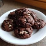 Stapel von dunklen, schokoladigen Easy Cookies And Cream Cookies mit wei&szlig;en St&uuml;ckchen auf einem wei&szlig;en Teller.