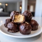 Ein Haufen Easy No Bake Peanut Butter Cheesecake Balls, einer davon angeschnitten, um die cremige Erdnussbutterf&uuml;llung zu zeigen.