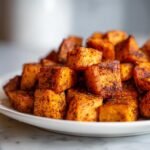 Nahaufnahme von gebratenen, gew&uuml;rzten S&uuml;&szlig;kartoffel-W&uuml;rfeln, die auf einem wei&szlig;en Teller gestapelt sind.