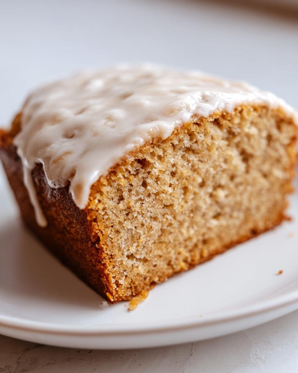 Nahaufnahme eines St&uuml;cks Rebel Float Frosted Banana Bread mit cremigem, wei&szlig;em Zuckerguss auf einem wei&szlig;en Teller.
